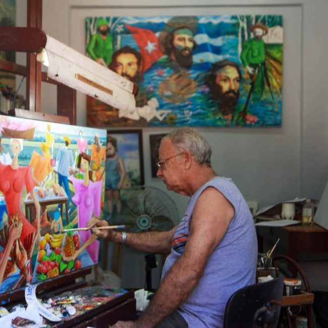 Senior artist working on colorful painting in indoor studio surrounded by vibrant artworks.