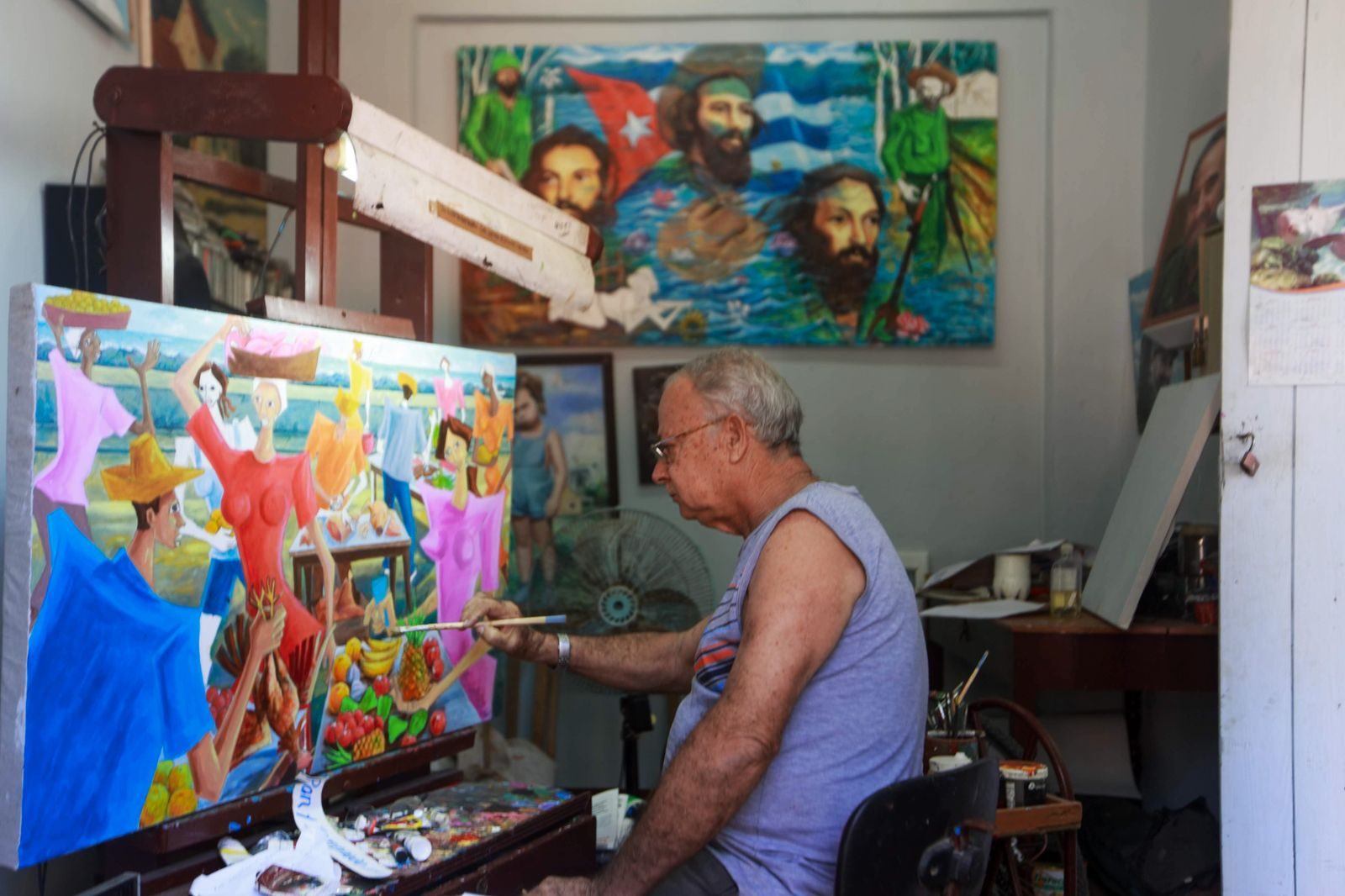 Senior artist working on colorful painting in indoor studio surrounded by vibrant artworks.